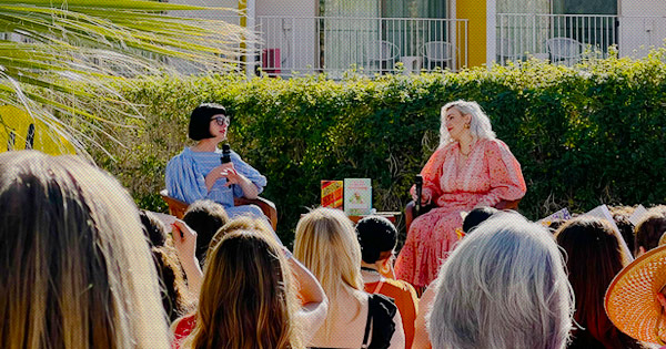 Two women sit on a stage. Two books are propped up on a table between them. The backs of women's heads are in the foreground watching the speakers. In the background are colorful hotel room balconies.