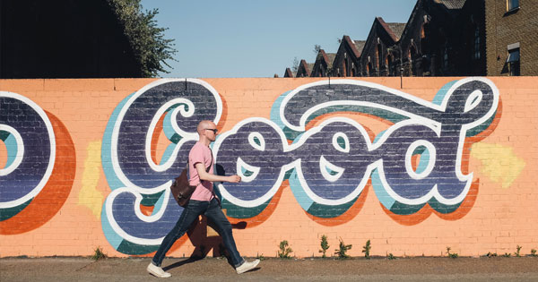 A man with a crossover bag walks past a wall with a mural that says Good.