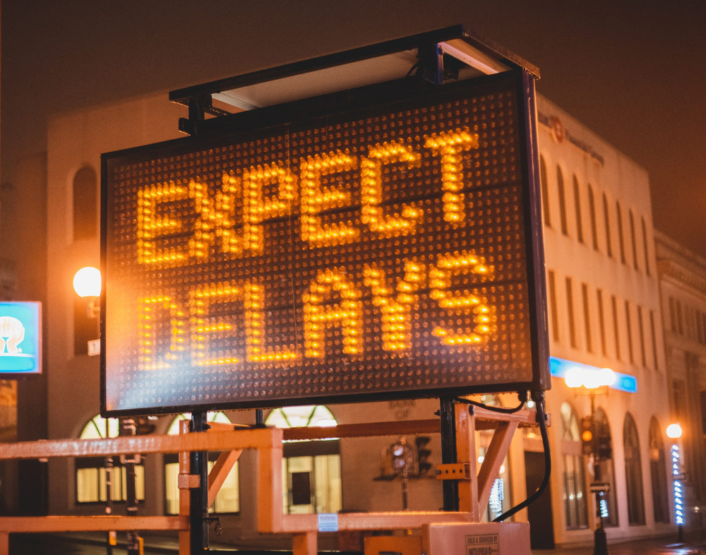 Construction roadwork illuminated sign that says Expect Delays. Background is a business area of a city. It is nighttime.