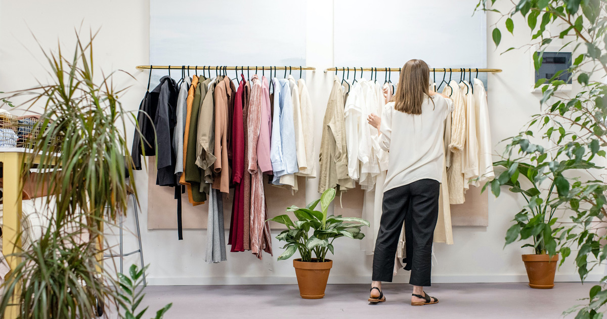 A woman standing with her back to the camera while she looks through a rack of clothing at a shop. Plants are on both sides off the photo and a plant sits just below the rack of clothing.