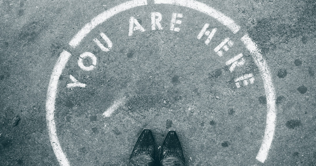 The photo is taken looking down at the ground. A white spraypainted stenciled circle and letters on asphalt reads You arrer here. The tips of shoes are shown at the edge of the photo standing within the circle.