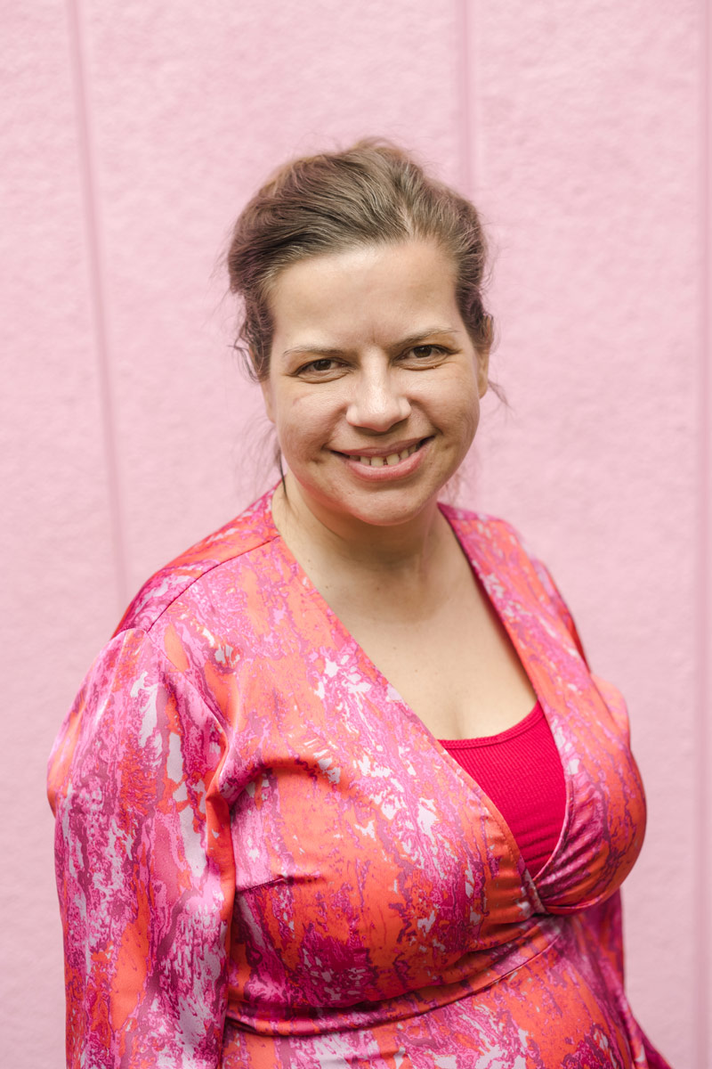 A woman Lisa Burford smiles at the camera with a pink wall behind her. She is wearing a pink multi-colored long sleeve dress. Her hair is in a messy ponytail.