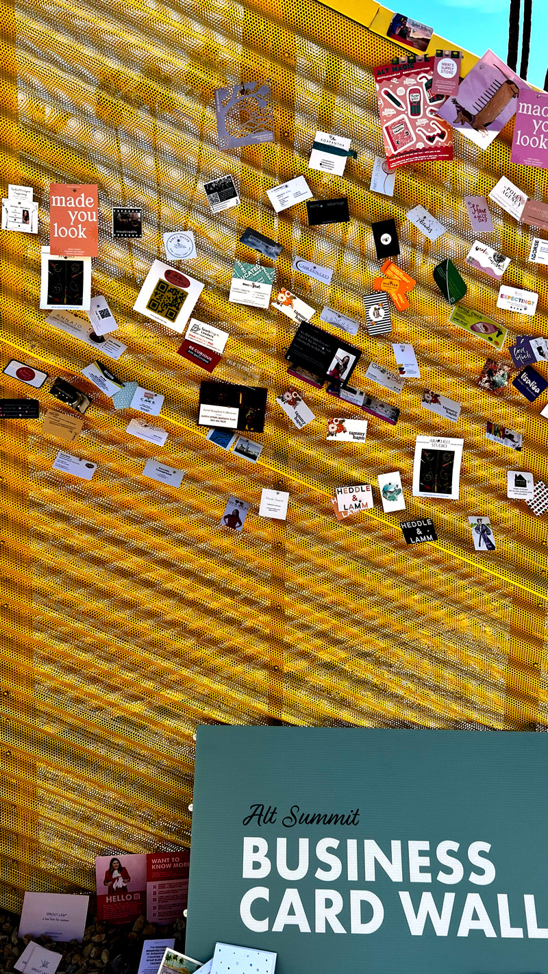 A metal mesh-like curvy wall with many business card designs of varying shapes and sizes attached to it stands behind a poster sign that reads Alt Summit Business Card Wall.