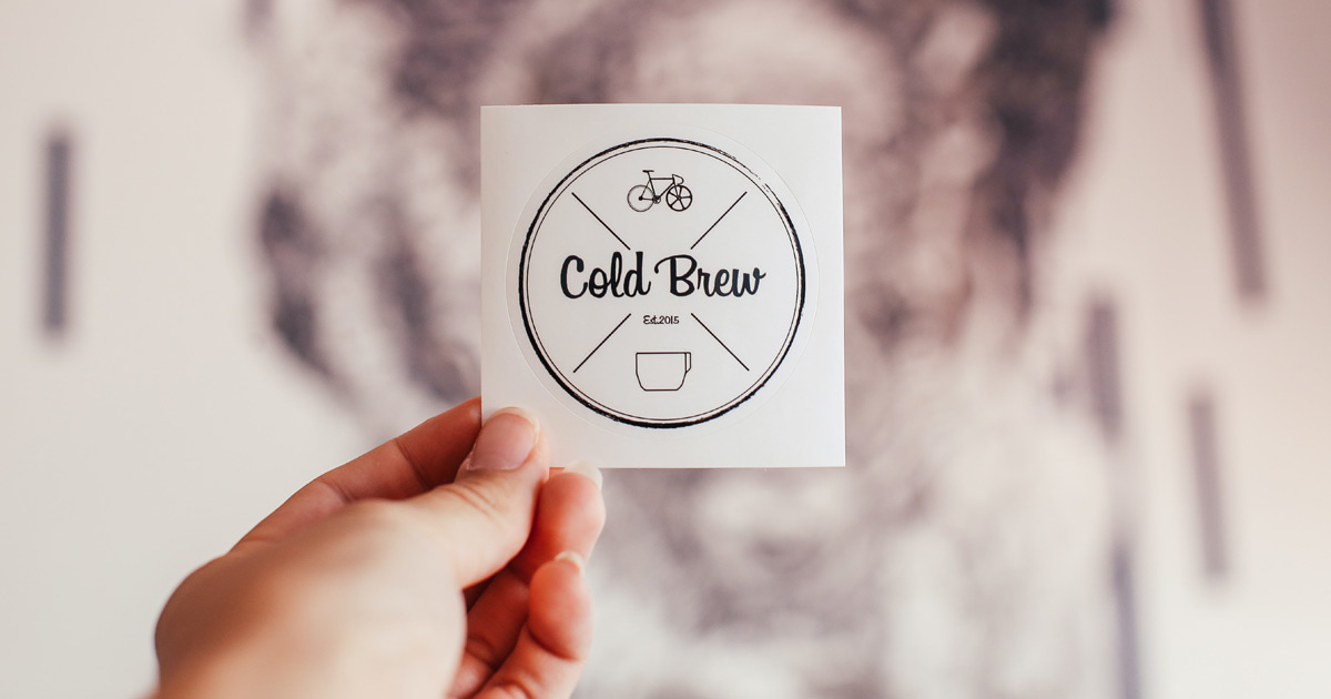 A hand holds up a round sticker with a logo on it. The logo reads Cold Brew Established 2015 and has icons of a bicycle and a coffee cup.
