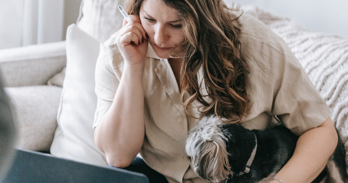 A woman sits on a couch with a small dog under her arm. She is leaning over staring at a laptop that is on a glass coffee table in front of her. Her face has a look of worry on it.