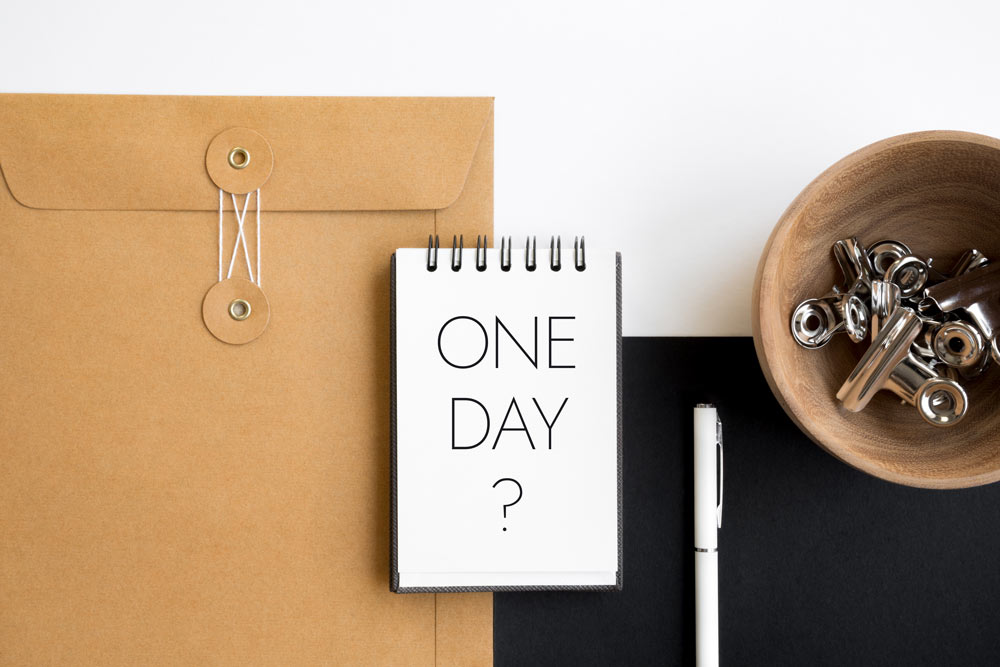 A notebook that says One Day? on top of a manilla envelope and black folder