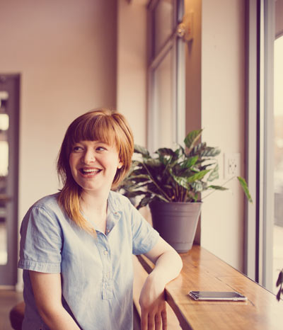 Woman with red hair smiling while sitting in front of a window with her phone on a table