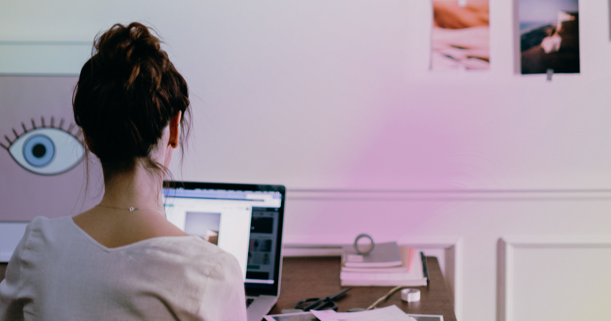 A woman with her hair in a messy bun sits at a desk looking at a laptop. There are images and photos pinned to the wall in front of her. Text on the image reads The Difference Between Web Designers and Web Developers