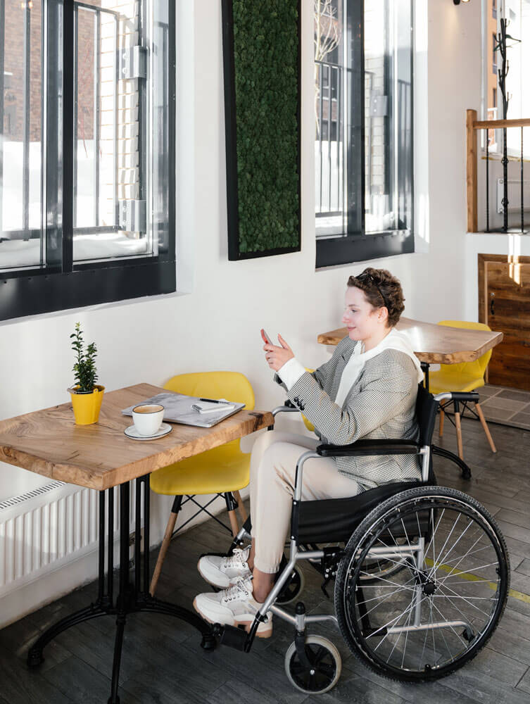 A person in a wheelchair in front of a coffeeshop table looking at their phone and smiling.