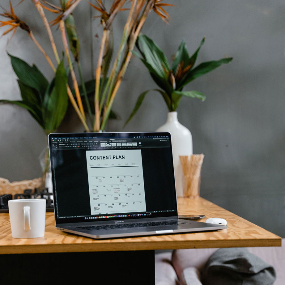 A laptop open on a table with a document on the screen that says Content Plan. A mug, earbuds case, phone, and plants are also on the table.