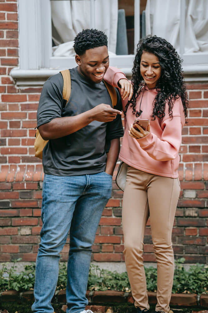 Two people standing outside looking at a phone a woman has in her hand. The man is pointing to the phone and they are both smiling.