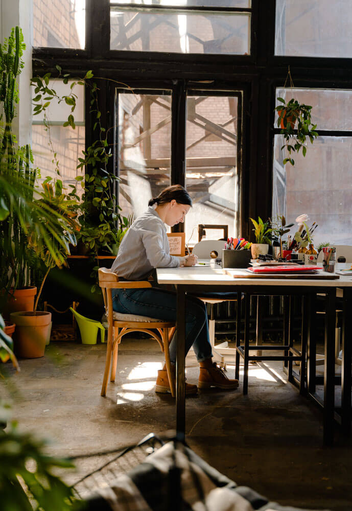 Person at a large studio table writing with a pen or pencil. On the table are different art supplies, plants, papers, and containers. There are very large windows along the wall behind them and many large plants fill the surrounding space.