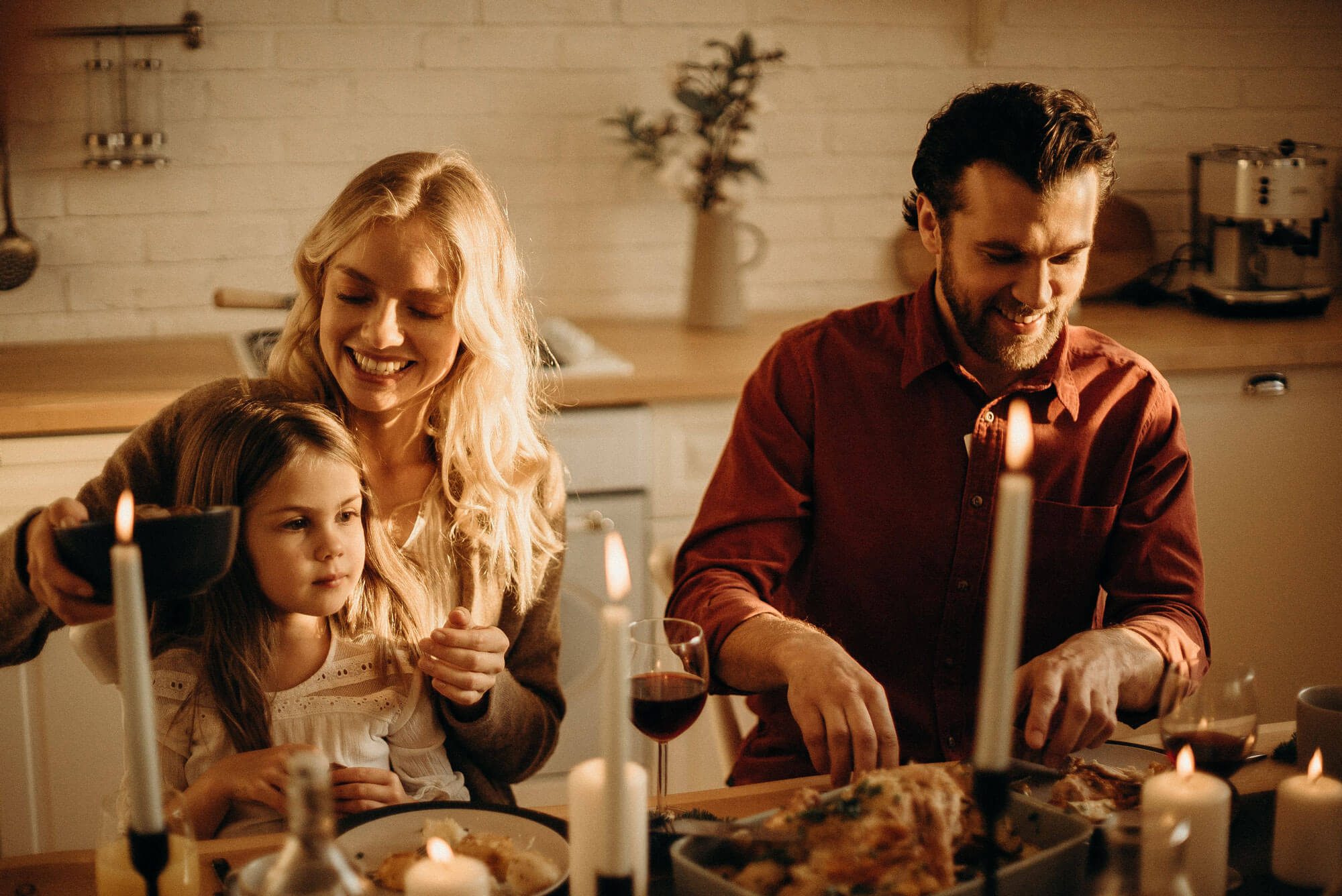 A family sitting at a table enjoying a holiday meal. There are candles lit on the table. A man and woman are smiling and a little girl is looking at the table of food in wonderment.
