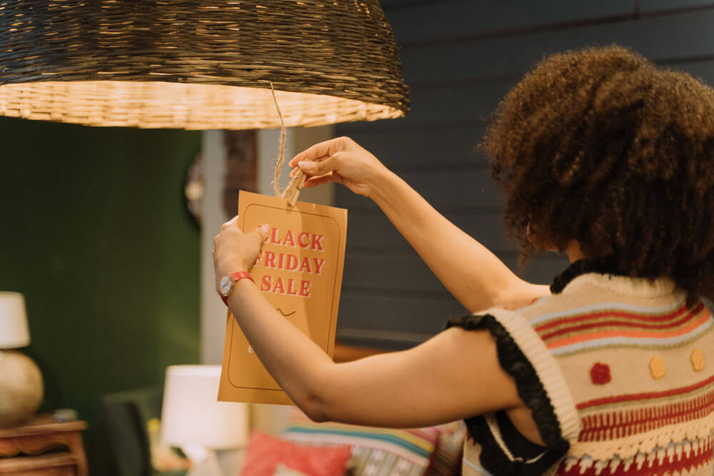 A person clipping a Black Friday Sale sign to a hanging lamp. Different products are in the backgroud by they are blurry.