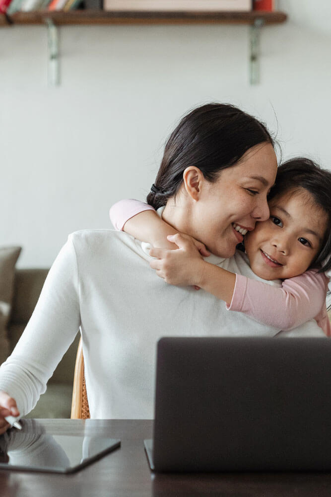 A mother sitting at a table with a laptop open in front of her and an iPad next to it. A young smiling girl is embrasing her from behind her back and the mom is turned townard her smiling.