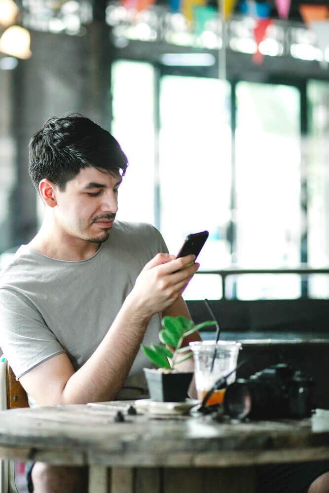 A person leaning back on a chair in a cafe looking at a phone they are holding in their hand. There is an iced beverage, a small plant, and a camera sitting on the table in front of him