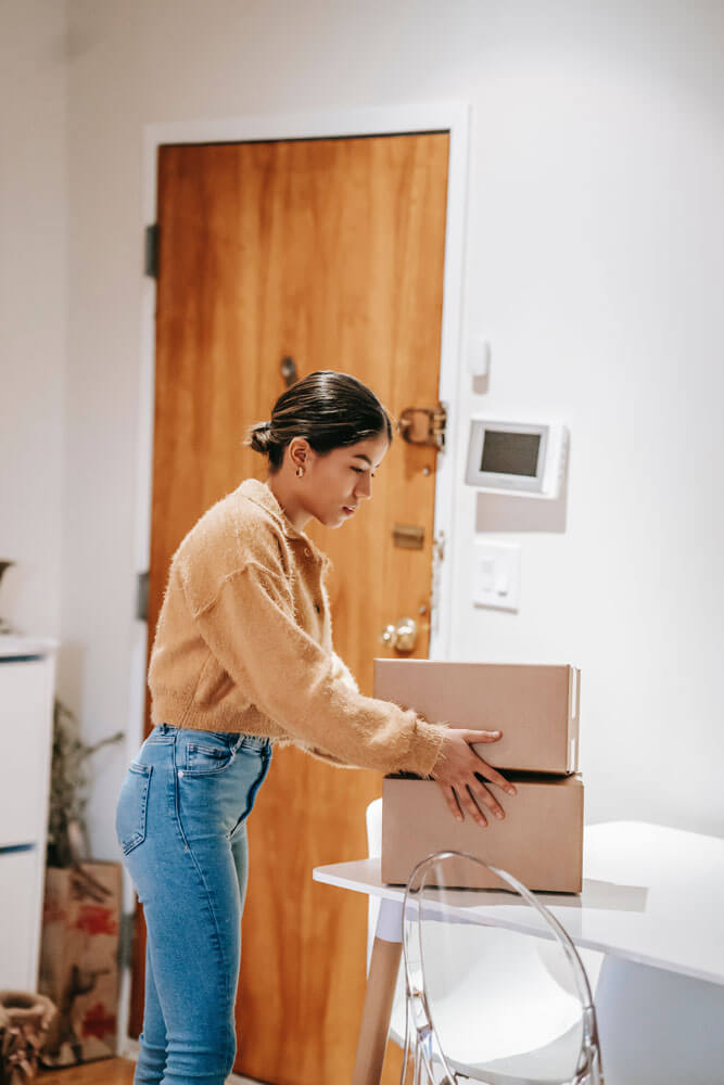 A woman with a ponytail holds two cardboard boxes that she's resting onto a table in front of her. The background room is a backroom of a business.